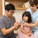武蔵小杉の「森のこどもクリニック小児科・皮膚科」にて、院長が幼児の右腕に予防接種を行っている様子。注射の痛みを和らげるため、看護師が接種部位のすぐ上にテントウムシ型の医療用機器「Buzzy Mini（バジーミニ）」を優しく当て、お子さんが落ち着いて受診できるようサポートしています。
